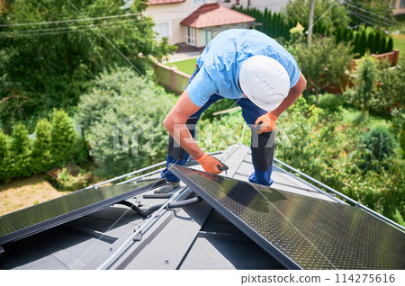 Worker building photovoltaic solar panel system on rooftop of house. Man engineer in helmets and gloves installing solar module with help of hex key outdoors. Renewable energy generation concept. Worker building photovoltaic solar panel system on rooftop of house. Man engineer in helmets and gloves installing solar module with help of hex key outdoors. Renewable energy generation concept. 114275616