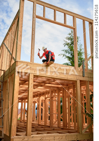 Wooden two-story frame house being built by carpenter. Man wearing glasses hammering nails using hammer, dressed in protective helmet and construction vest. Concept of modern ecological construction. Wooden two-story frame house being built by carpenter. Man wearing glasses hammering nails using hammer, dressed in protective helmet and construction vest. Concept of modern ecological construction. 114275618