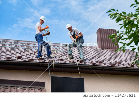 Men workers installing solar panel system on roof of house. Electricians in helmets lifting up photovoltaic solar module with help of ropes outdoors. Concept of alternative and renewable energy. Men workers installing solar panel system on roof of house. Electricians in helmets lifting up photovoltaic solar module with help of ropes outdoors. Concept of alternative and renewable energy. 114275621