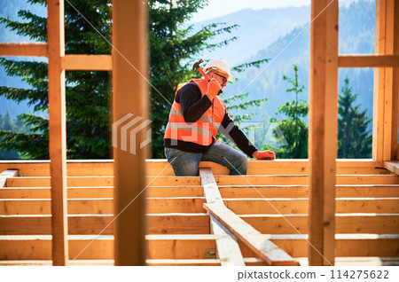 Carpenter constructing two-story wooden frame house near forest. Man hammering nails into the structure, wearing protective helmet and construction vest. Concept of modern ecological construction. Carpenter constructing two-story wooden frame house near forest. Man hammering nails into the structure, wearing protective helmet and construction vest. Concept of modern ecological construction. 114275622