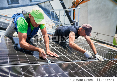 Workers building photovoltaic solar panel system on rooftop of house. Men engineers installing solar module with help of wrench outdoors. Alternative and renewable energy generation concept. 114275624
