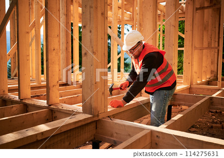 Carpenter constructing wooden frame house. Bearded man with glasses is hammering nails into the structure, wearing protective helmet and construction vest. Concept of modern ecological construction. Carpenter constructing wooden frame house. Bearded man with glasses is hammering nails into the structure, wearing protective helmet and construction vest. Concept of modern ecological construction. 114275631