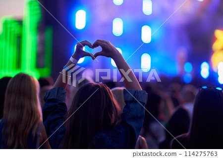 Person forms heart shape with hands at lively outdoor music festival, blurred stage lights, crowd enjoys event. Love symbol gesture, fans celebrate artist, youth culture, happy summer vibe in concert. Person forms heart shape with hands at lively outdoor music festival, blurred stage lights, crowd enjoys event. Love symbol gesture, fans celebrate artist, youth culture, happy summer vibe in concert. 114275763