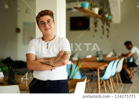 Confident transgender pro in casual office attire stands with arms crossed. Warm, welcoming smile in creative co-working space. Inclusive workplace environment, gender diversity embraced. 114275771