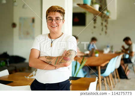 Confident transgender male employee smiling in office space, arms crossed, diverse team working in background. Inclusion at modern workplace, LGBTQ representative 114275773