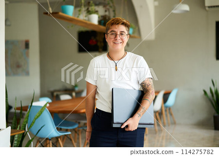 Confident transgender person with tattoos holds laptop, smiles in modern eco office. Inclusive LGBTQ work environment, gender identity acceptance at tech startup. Casual corporate attire. Confident transgender person with tattoos holds laptop, smiles in modern eco office. Inclusive LGBTQ work environment, gender identity acceptance at tech startup. Casual corporate attire. 114275782