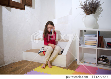 Elementary age school girl talking on smart mobile phone, expressing surprised emotion, sitting on steps at home, nearby a bookshelf with books Elementary age school girl talking on smart mobile phone, expressing surprised emotion, sitting on steps at home, nearby a bookshelf with books 114277844