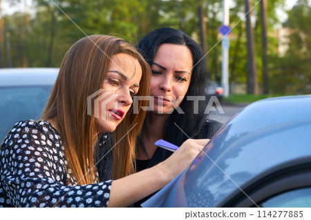 Young woman with her friend signing a document on the trunk of a car Young woman with her friend signing a document on the trunk of a car 114277885