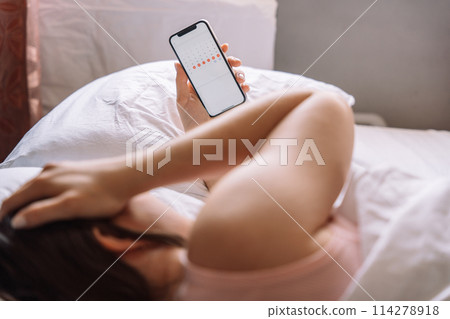 The girl has stomach pain and lies in bed holding a phone with a calendar in her hand 114278918