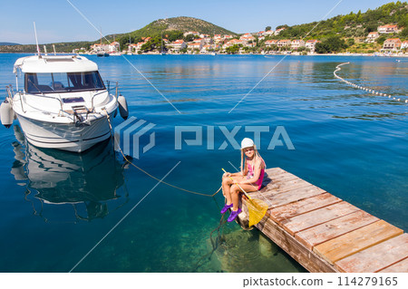 Cute little girl kid sits on wooden pier next moored boat yacht with scenic turquoise sea water background. Happy child in cap hold fishing net have fun explore nature at summer vacation 114279165