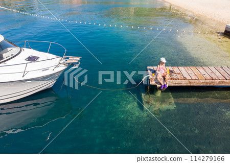 Cute little girl kid sits on wooden pier next moored boat yacht with scenic turquoise sea water background. Happy child in cap hold fishing net have fun explore nature at summer vacation 114279166