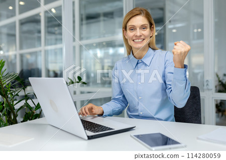 Successful woman looking at camera with toothy smile and making winner gesture in front of table with pc. Caucasian female in formal clothes clenching fist with pleasure from career achievement. 114280059