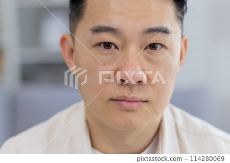 A detailed close-up portrays a middle-aged Asian man with a contemplative, serious expression, captured indoors with a soft focus background. A detailed close-up portrays a middle-aged Asian man with a contemplative, serious expression, captured indoors with a soft focus background. 114280069