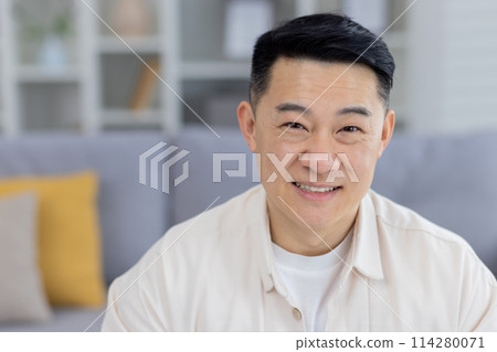 Portrait of a happy middle-aged Asian man sitting in a contemporary living room setting, wearing a casual cream shirt with a relaxed, confident smile. 114280071