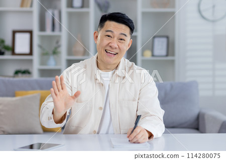 Cheerful Asian man sitting at home, talking and waving to the camera during an online meeting. He showcases a friendly and welcoming demeanor. 114280075