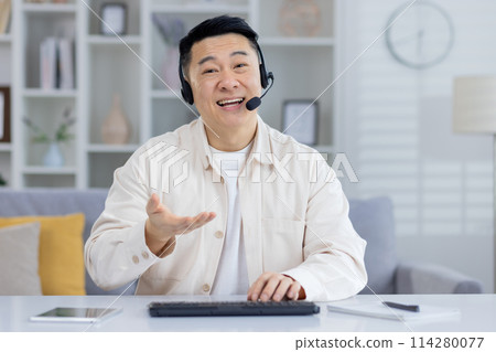 Cheerful Asian male in a beige shirt using a headset in a home office, looking at camera with a smile, possibly in a customer service call. Cheerful Asian male in a beige shirt using a headset in a home office, looking at camera with a smile, possibly in a customer service call. 114280077