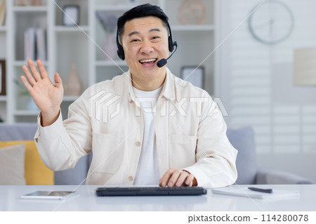 Cheerful Asian man waving during a video conference at home. He sits in a well-lit room, showcasing a professional yet relaxed atmosphere. Cheerful Asian man waving during a video conference at home. He sits in a well-lit room, showcasing a professional yet relaxed atmosphere. 114280078