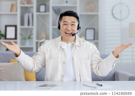 Happy middle-aged Asian man wearing headset, smiling and gesturing while providing customer service from a cozy home office setup. 114280089