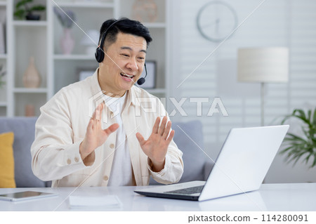 A joyful older Asian man wearing a headset and interacting cheerfully with someone on his laptop from a well-decorated living room. A joyful older Asian man wearing a headset and interacting cheerfully with someone on his laptop from a well-decorated living room. 114280091