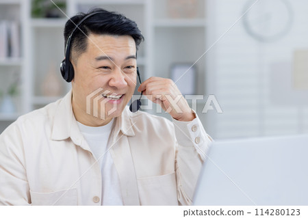 A cheerful Asian man wearing a headset smiles while working from his home office. He is interacting and providing customer support with positivity and professionalism. A cheerful Asian man wearing a headset smiles while working from his home office. He is interacting and providing customer support with positivity and professionalism. 114280123