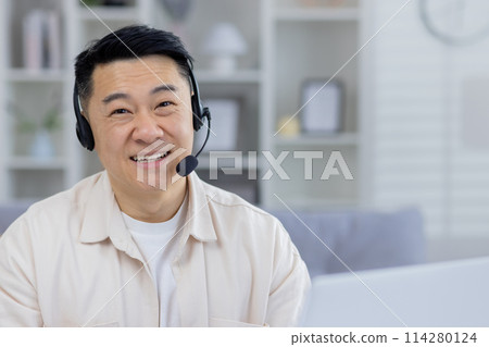 Friendly Asian male professional using a headset in a modern home office setting, looking cheerful and attentive while engaging in customer service or telecommuting. Friendly Asian male professional using a headset in a modern home office setting, looking cheerful and attentive while engaging in customer service or telecommuting. 114280124