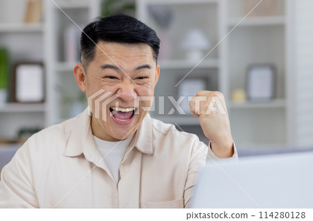 Joyful middle-aged Asian man in a casual shirt celebrating a triumph in front of his laptop, expressing excitement and happiness in a home office setting. 114280128