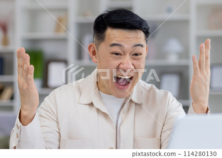 A middle-aged Asian man shows great excitement and joy while working at his computer in a modern home office setting. His exhilarated expression suggests a moment of triumph. 114280130