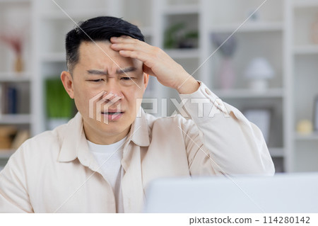 A middle-aged Asian man looking stressed with a hand on his forehead, sitting at a white desk in a brightly lit office environment, showing signs of work-related stress and exhaustion. 114280142