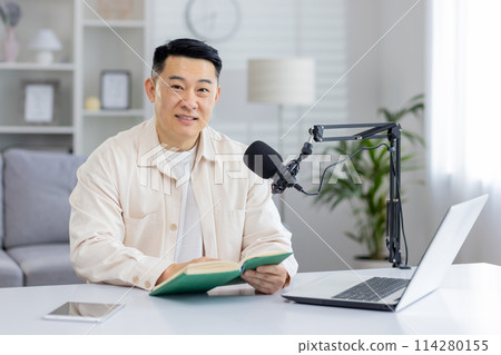 A focused man in a stylish, bright home office setup engrossed in a book with a podcasting microphone mounted on the desk. 114280155