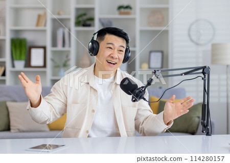 Enthusiastic Asian podcaster talking with hands, having an engaging conversation indoors, looking at camera with headphones and microphone. 114280157