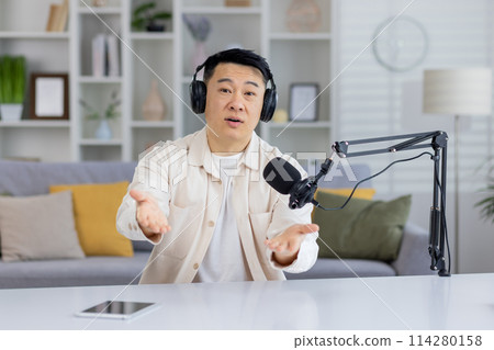 An engaging Asian man talking and looking at the camera while recording a podcast in a home studio setting. 114280158