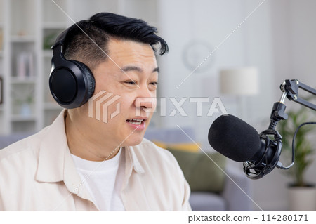 Asian male podcaster speaking into a microphone while recording content for his podcast in a cozy home studio setting. Asian male podcaster speaking into a microphone while recording content for his podcast in a cozy home studio setting. 114280171