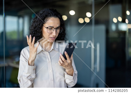 Upset thinking business woman close up inside office, young hispanic woman reading bad news on phone online, business woman at workplace holding smartphone typing text message 114280205