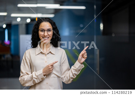 A professional businesswoman with curly hair smiles and points beside her in a modern office setting, exuding confidence and positivity. A professional businesswoman with curly hair smiles and points beside her in a modern office setting, exuding confidence and positivity. 114280219