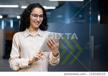 A joyful professional woman in smart attire, using a digital tablet in a modern office setting, reflecting productivity and connectivity. 114280220