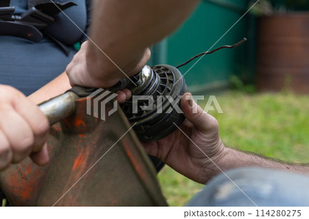 Replacing the nylon thread on a grass trimmer. A man unscrews the nut to change the line on the head of a trimmer while mowing the grass. Grass trimmer concept. Replacing the nylon thread on a grass trimmer. A man unscrews the nut to change the line on the head of a trimmer while mowing the grass. Grass trimmer concept. 114280275