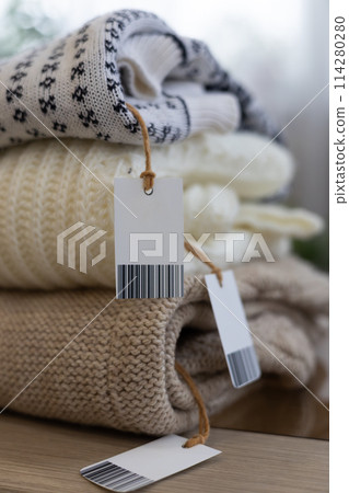 Close-up of folded clothes with tags and space for writing on them. Selling unwanted clothes from your wardrobe for use. A neat stack of wool clothes is folded on a table in the living room. 114280280