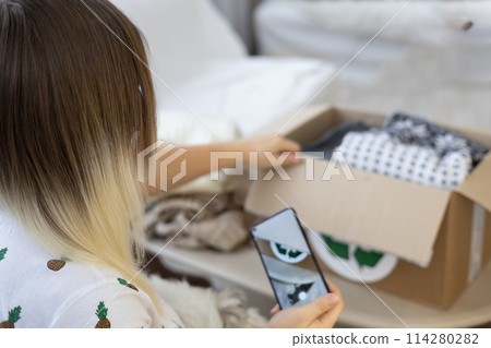 Having put old clothes in a box, a woman takes a photo of it on her phone so that she can post an advertisement for sale on the Internet. Sorting clothes and selling them online. Recycling clothing. 114280282