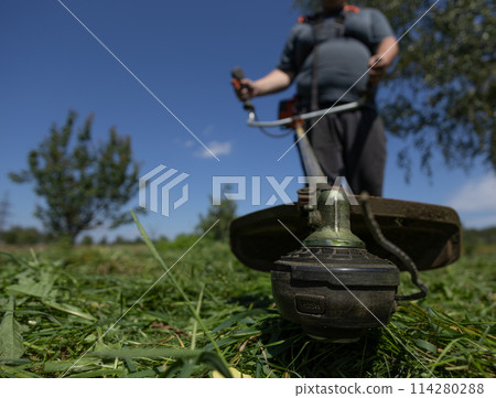 Wide viewing angle. A man with a trimmer mows the grass on the lawn behind the house on a sunny summer day. A man holds a trimmer in front of him and mows thick, lush grass. Wide viewing angle. A man with a trimmer mows the grass on the lawn behind the house on a sunny summer day. A man holds a trimmer in front of him and mows thick, lush grass. 114280288