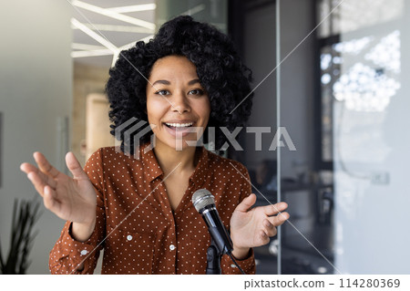 Confident young African American woman speaking passionately at a business seminar or conference, holding a microphone, in a modern office setting. Confident young African American woman speaking passionately at a business seminar or conference, holding a microphone, in a modern office setting. 114280369
