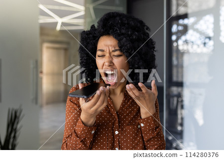 African American woman in a polka dot shirt expressing anger and frustration while yelling into a mobile phone in a modern office setting. 114280376