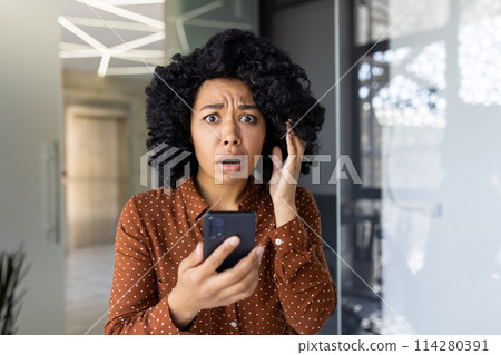 A young woman in a polka dot shirt looks shocked and upset while reading bad news on her smartphone in a modern office setting. 114280391