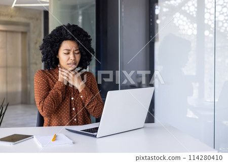 A young African American woman holding her throat, feeling unwell with a sore throat as she works at her desk in a modern office setting. A young African American woman holding her throat, feeling unwell with a sore throat as she works at her desk in a modern office setting. 114280410