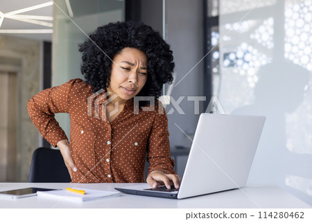 A young African American woman grimaces in pain, holding her lower back while working at her laptop in a bright modern office. 114280462