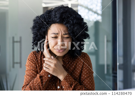 An African American woman experiences discomfort, holding her ear in pain, possibly suffering from otitis or an earache, depicted in a workplace setting. 114280470