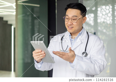 Smiling Asian male doctor in a white coat holds a tablet, engaging in a video call with a patient or colleague. Modern healthcare communication shown in an office setting. Smiling Asian male doctor in a white coat holds a tablet, engaging in a video call with a patient or colleague. Modern healthcare communication shown in an office setting. 114281516