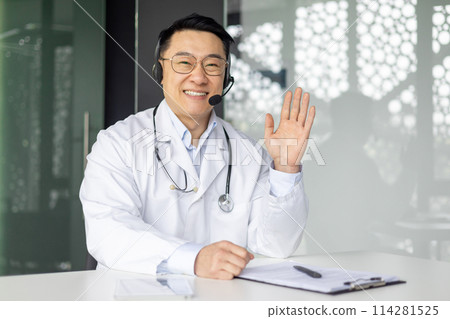 A cheerful Asian male doctor in a lab coat and headset waves hello during a telehealth consultation, sitting at his desk in a modern clinic. A cheerful Asian male doctor in a lab coat and headset waves hello during a telehealth consultation, sitting at his desk in a modern clinic. 114281525