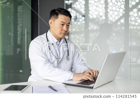 A focused Asian mature male doctor wearing a lab coat and stethoscope is working intently on a laptop in a well-lit modern office environment. A focused Asian mature male doctor wearing a lab coat and stethoscope is working intently on a laptop in a well-lit modern office environment. 114281545