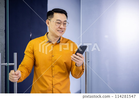 Cheerful Asian businessman in a vibrant orange shirt using a smartphone near office doors. He looks content and connected, standing in a modern environment. Cheerful Asian businessman in a vibrant orange shirt using a smartphone near office doors. He looks content and connected, standing in a modern environment. 114281547
