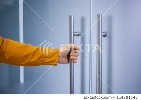 Close-up of a person's hand gripping a metallic handle to open a glass door, emphasizing themes of access, entry, or beginning a new opportunity. Close-up of a person's hand gripping a metallic handle to open a glass door, emphasizing themes of access, entry, or beginning a new opportunity. 114281548